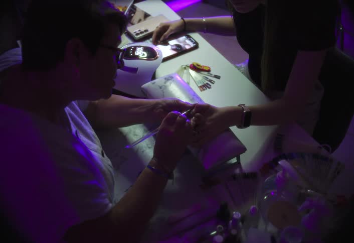 A nail technician in the foreground is attentively working on a clients hand using a small brush to apply material to a fingernail with the surrounding white table covered in tools and equipment