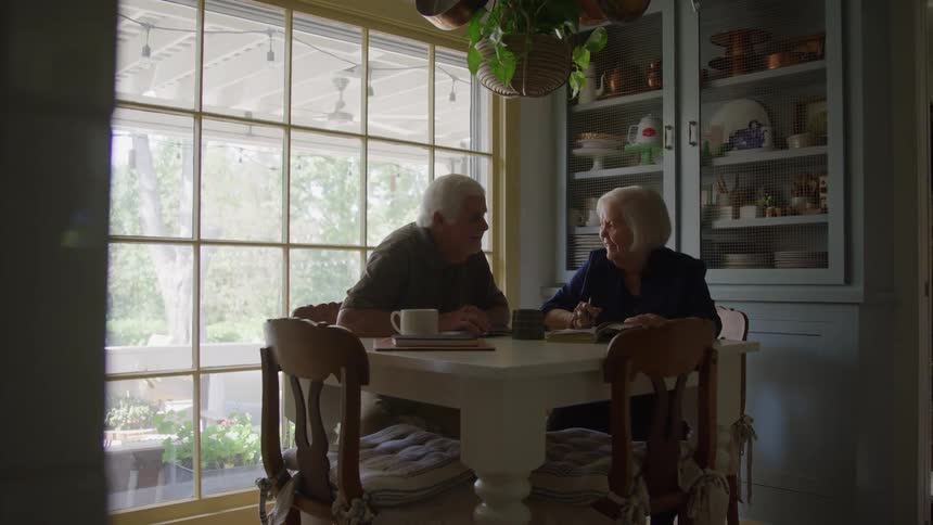 An older man and woman are sitting at a small white kitchen table talking and smiling across from each other in the morning light with a large window and a blue hutch in the background