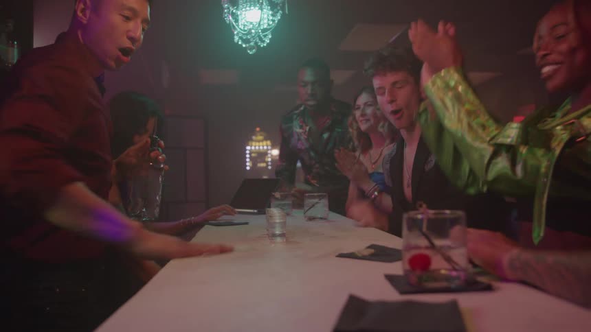 A lively group of five men and three women in stylish club attire sit around a white table in a dimly lit club with neon lights and a chandelier enjoying drinks and cheering happily
