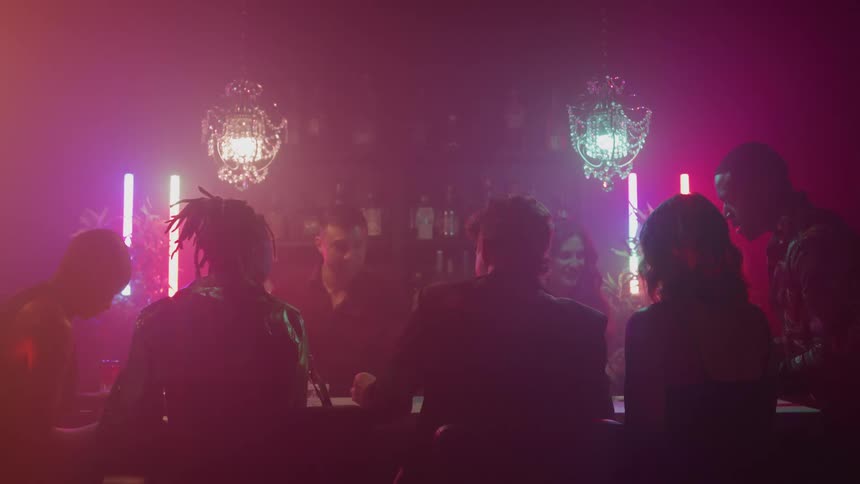 A group of people is sitting together at a table in a dimly lit atmospheric bar or club the background is illuminated by decorative chandeliers and vertical neon tube lighting casting warm and colorful hues
