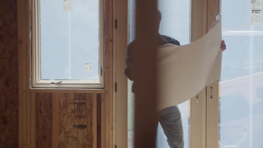 A tall smiling older worker wearing a plaid shirt and jeans stands by a glass door inside a house under construction holding a large piece of architectural paper and reading the plans
