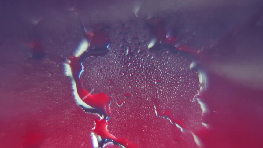 Tiny water droplets form a patch of condensation on a wet glass surface with larger drops streaking down against a blurry red and purple background