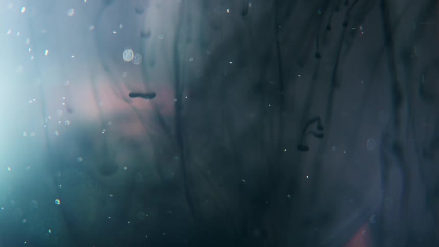 The video is an abstract closeup of dark ink spreading and dissolving in a pale fluid mediumbright particles or bubbles drift throughout the frame with subtle pink and cyan highlights suggesting light refraction