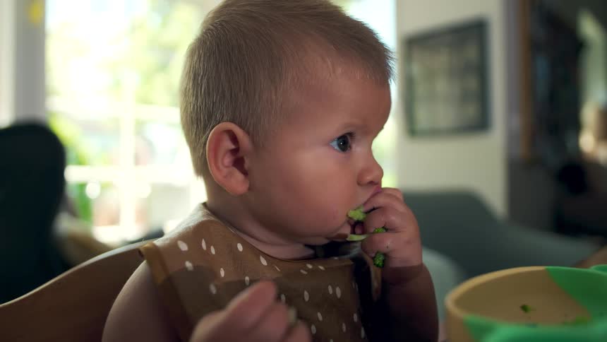 An infant eating with her hands putting food into her mouth while sitting at the dining table