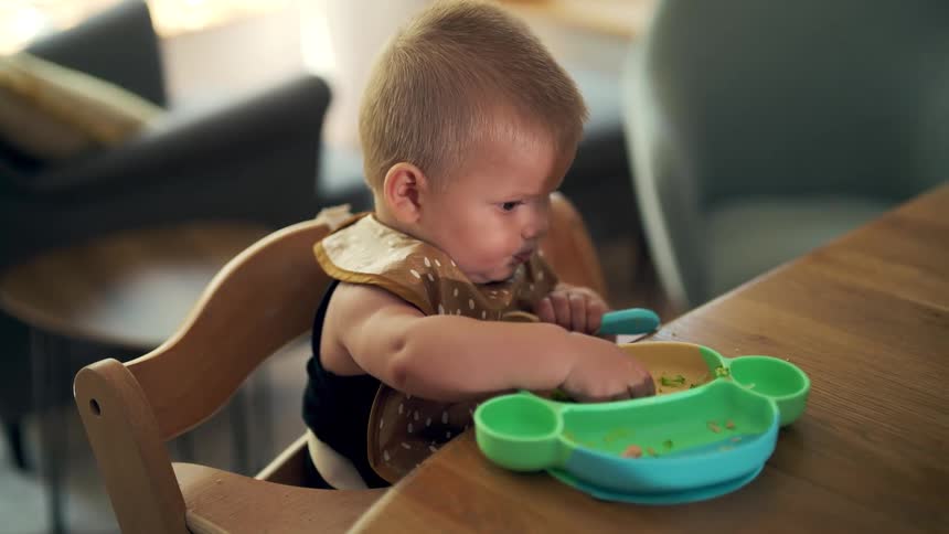 A little blond boy wearing a bib is sitting in a wooden chair and eating from a teddy bearshaped object