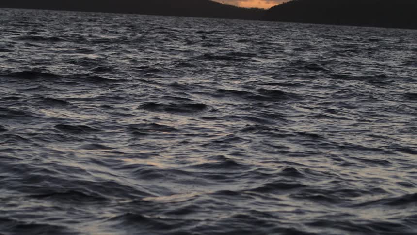 A slowmotion shot begins on dark choppy water and tilts up to reveal a distant island silhouetted against a beautiful golden sunset