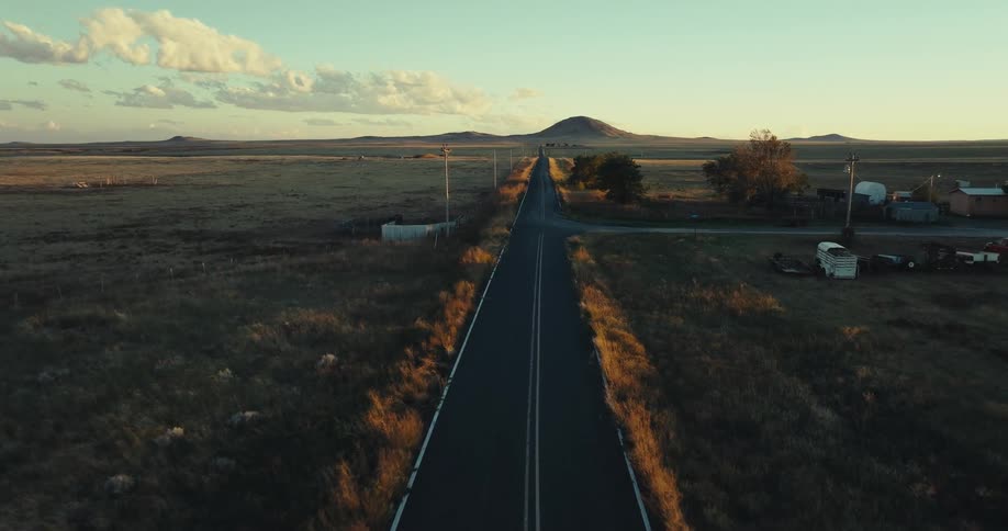 The lone straight highway stretches into the fading light its white lane dividers converging on the distant horizon on both sides the vast flat fields of green and brown farmland recede under a pale evening sky