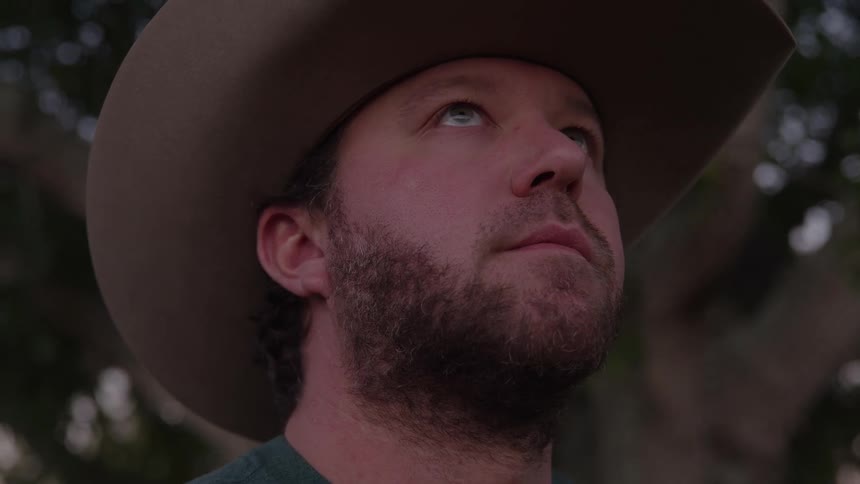 A bearded man in a dark widebrimmed cowboy hat stands outdoors tilting his head slightly as he looks intently upward the low angle emphasizes his silhouette against the bright sky and the shadow cast over his face giving him a thoughtful and rugged a