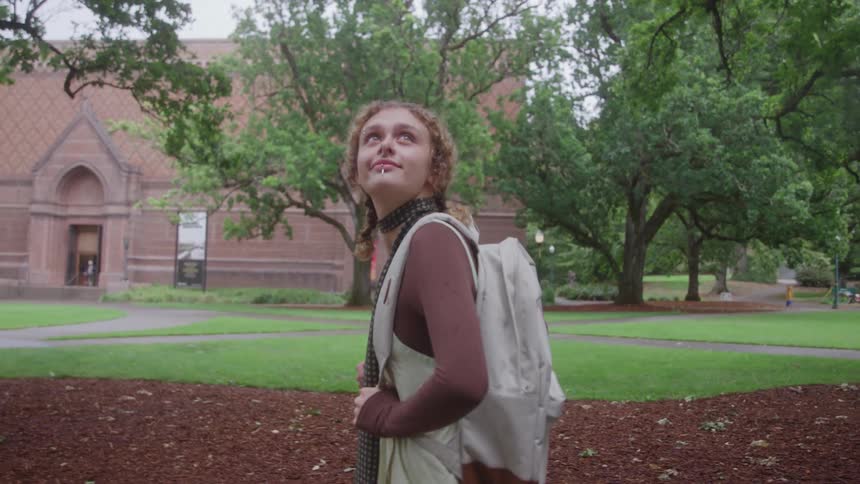 A young woman with curly hair tied back and wearing a lightcolored backpack stands outdoors looking up and then turning her head she is on a mulched patch with a tree and in the background is a grassy lawn and a brick building suggesting a university