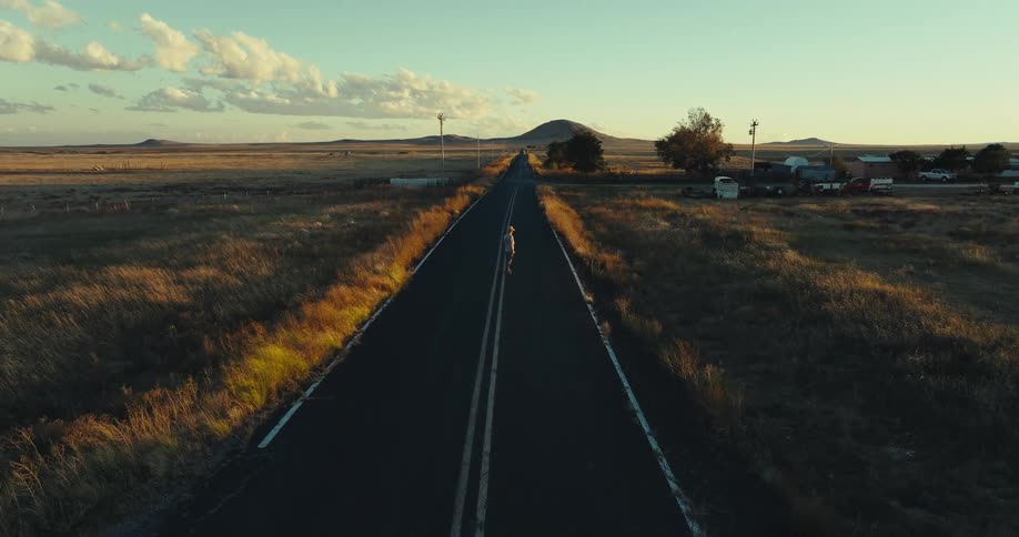 A man skateboarding on an empty road in a rural community with brown grass lining both sides of the road at sunset