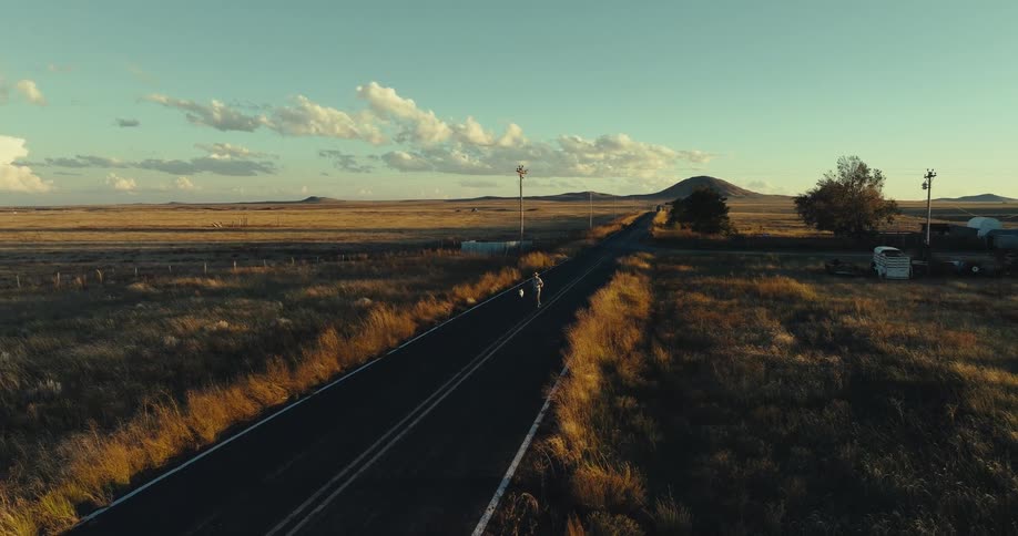 An elevated wide shot captures a person skateboarding along a narrow isolated country road that stretches through a dry grassy plain at sunset in the distance a distinct mesa or hill rises from the flat landscape under a blue sky with scattered cloud