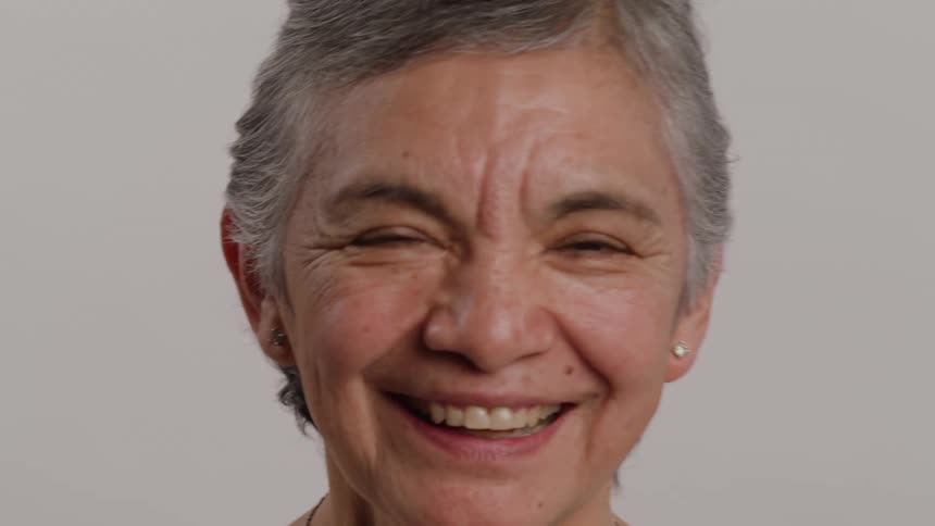 The video is a closeup shot of an older woman with short gray hair and earrings set against a plain backgroundshe has a wonderfully genuine and joyful expression smiling broadly with her mouth open