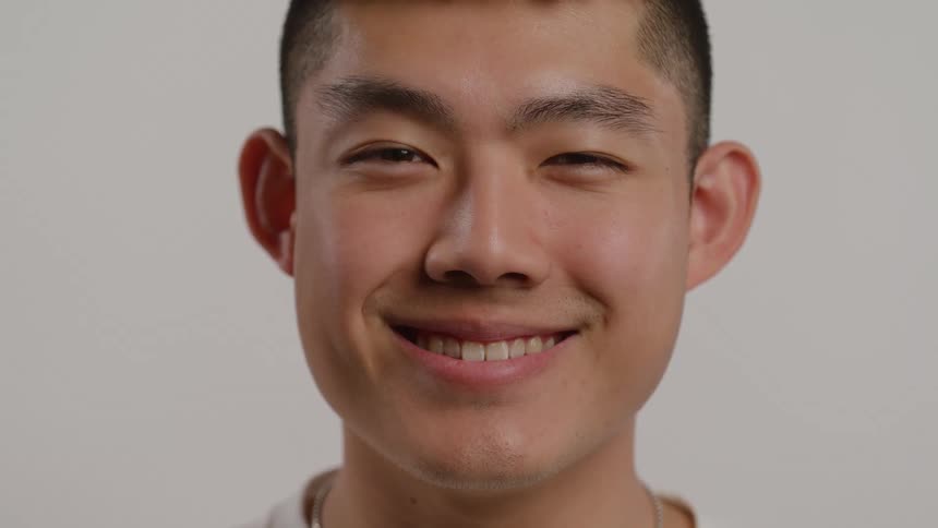 This video is a closeup portrait of a young asian man with a short buzz cut set against a plain light background he is smiling genuinely with his eyes slightly crinkled conveying a feeling of friendliness and contentment
