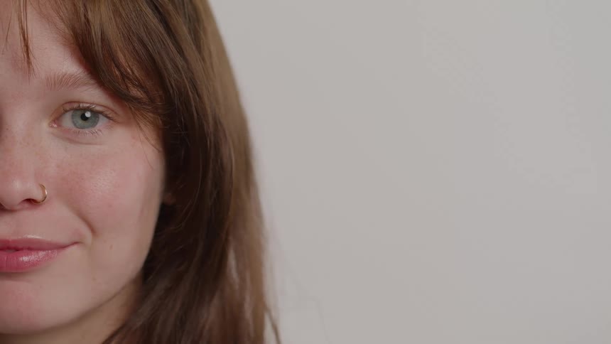 The camera holds on the right half of a smiling young womans face highlighting her bluegreen eye and a small nose ring her light brown hair and a scattering of freckles are visible against the plain bright background