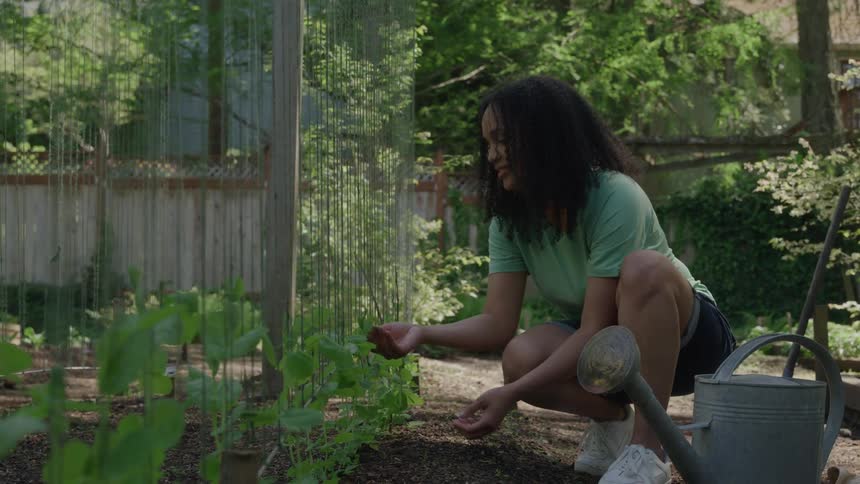 A woman with dark curly hair crouches down in a sundappled garden plot gently tending to a row of small leafy green plants she smiles as she works with a metal watering can resting nearby and a wooden fence and lush greenery forming the backyard bac
