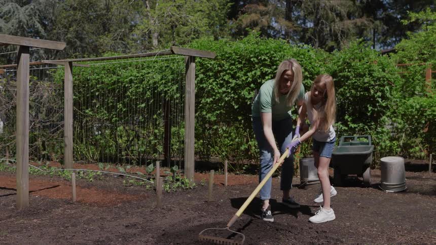 The video shows a woman and a young girl working together in a garden the woman is guiding the girl as they use a longhandled rake to smooth out and prepare the dark soil for planting