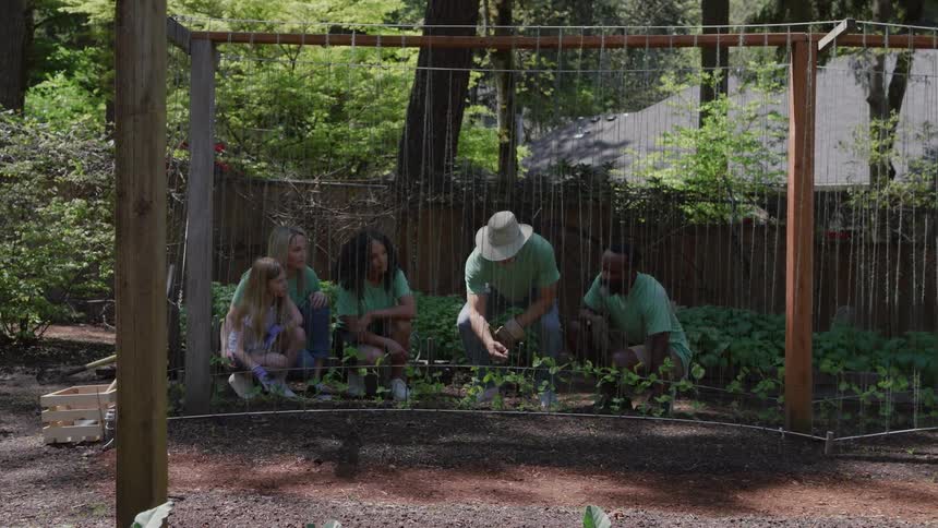 A group of five people gathered in a community garden or large yard tending to plants in a circular fencedin area there are three adults and two children one young girl and another person who appears to be a teenager or young adult all squatting dow