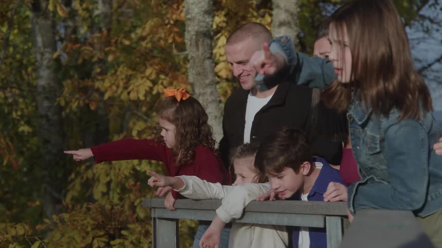 A family of six mother father and four children is gathered around a wooden railing on a bright autumn day pointing offcamera to the left capturing a shared moment of interest against a backdrop of fallcolored trees