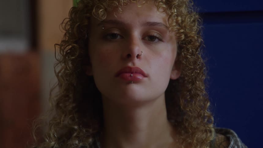A young woman with tight curly blonde and brown hair looking straight at the camera with a neutral expression prominently displaying multiple facial piercings