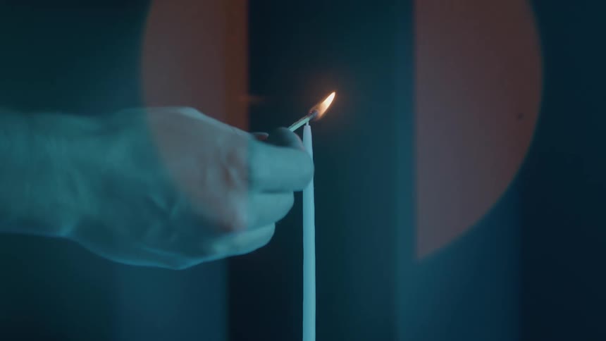 A closeup shot shows a hand holding a lit match near the wick of a tall thin white candle in a dimly lit bluetoned room the flame successfully transfers from the match to the candles wick lighting it