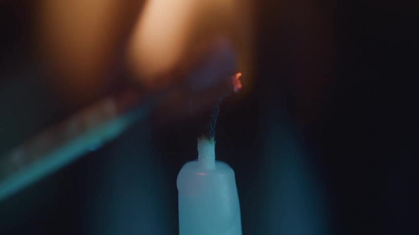 A match lights the candles wick producing a steady soft flame against a dark blurry background