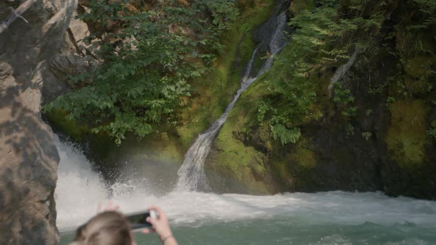 Woman in boat taking photographs with cell phone of waterfall
