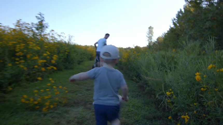 A toddler in a white cap runs in a flowerlined path toward an adult under a clear sky