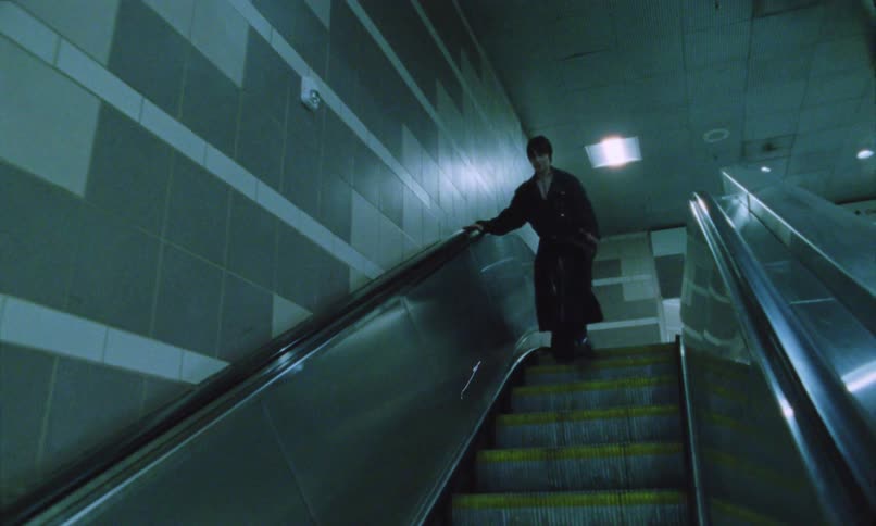 Two person wearing black clothing are seen walking quickly down an escalator in a subway setting with bluegreen tiled walls where the lighting is dim and casts a shadowy atmosphere