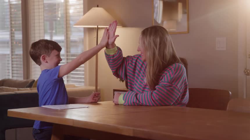 N adult woman and a young boy sitting at a wooden table likely doing homework or an activity together in a brightly lit homethe boy after completing something on paper raises his hand for a highfive which the woman enthusiastically returns showin