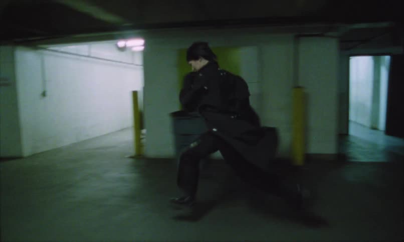 A young woman wearing a jacket runs inside an underground parking lot at night with several cars are parked