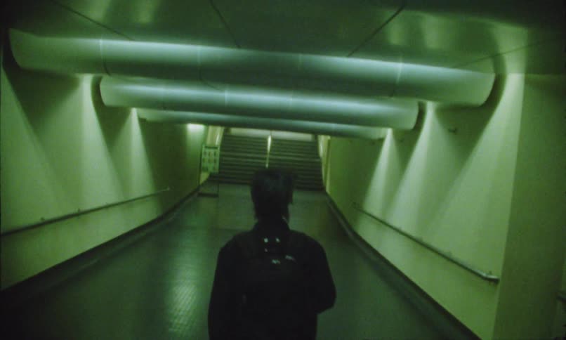 Back of a person wearing black clothes and carrying a backpack walking down a corridor with a staircase at the end indoors