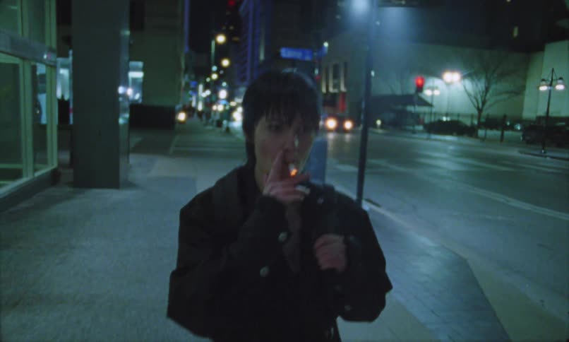 A shorthaired woman wearing dark clothes walks down a city sidewalk at night briefly stopping in her stride to light a cigarette near a streetlamp with the headlights of distant cars and the dark outlines of urban buildings forming the background
