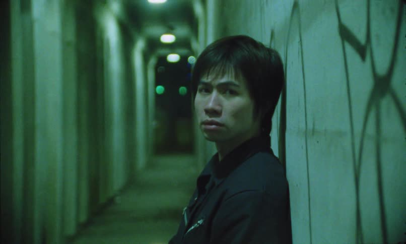 A young man smoking a cigarette rests against a wall in a hallway illuminated by ceiling lights at night