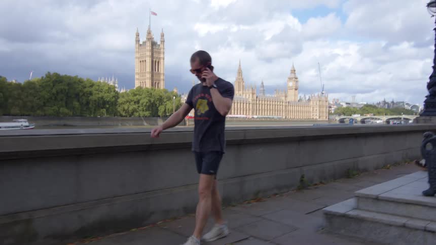 A man in dark casual clothes and sunglasses is walking along a paved riverside path while talking on a mobile phone with the historic towers and buildings visible across the river under a partly cloudy sky