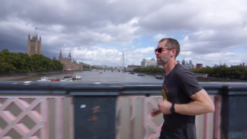 A man in sunglasses jogs across a bridge under a cloudy sky with londons iconic palace of westminster visible in the distance
