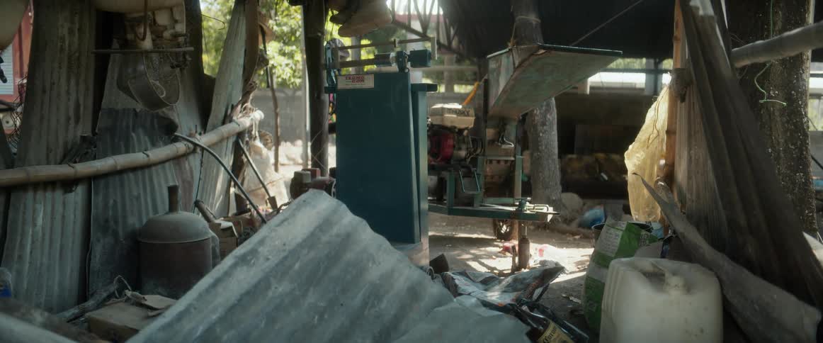 A cluttered openair workshop holds corrugated metal sheets jugs and aging machinery beneath a patchwork roof