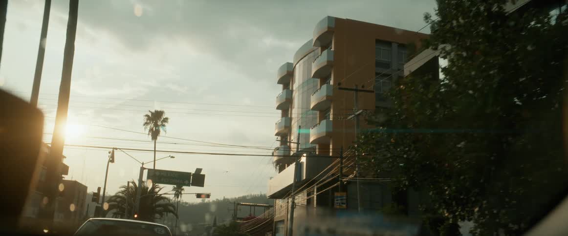 A car drives through a city street lined with modern buildings and trees at sunset