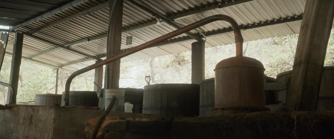 A rustic distillation setup with a long copper pipe sits in an opensided shed beside large barrels