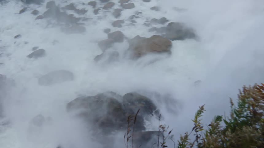 A vast amount of foamy white water dramatically tumbles over a rocky ledge while the camera pans from the falling water to focus briefly on the large wet boulders at the bottom framed by some dense green shrubbery on the right