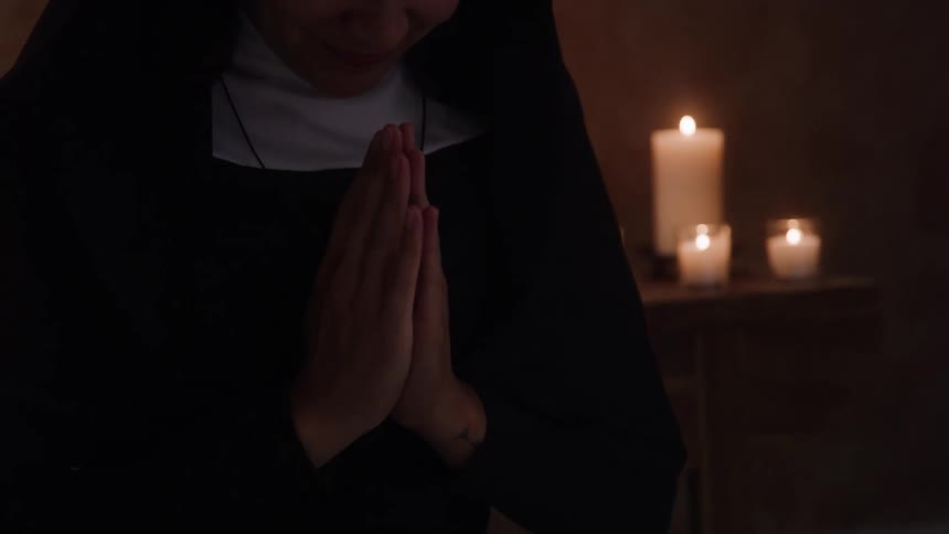 A nun prays in a dim room with candles glowing behind her