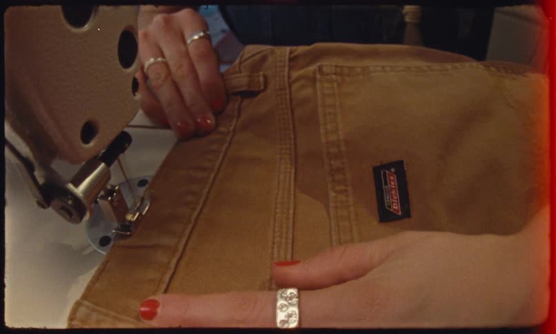 A person guides tan pants under a sewing machine their ringed hand holding the fabric steady