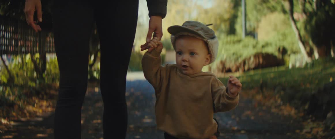 The video is a closeup of a toddler wearing a cap and a brown outfit happily taking clumsy steps while being led by an adult holding their handthey are walking together down a leafy sundappled path suggesting a pleasant stroll in a park or neighbo