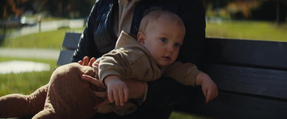 A little blond boy is sitting on the lap of a woman who is sitting on a public park bench and he is fidgeting restlessly