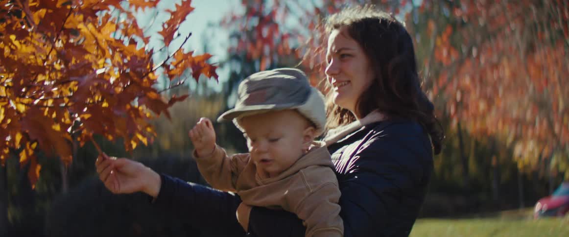 The woman holds the baby up to the fall leaves on the tree