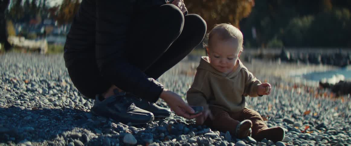 The video features a tender moment outdoors as an adult whose legs are visible in dark athletic wear crouches beside a toddler on a rocky grey beach the adult gently offers a small colorful stone to the baby who is focused on the stones scattered ar