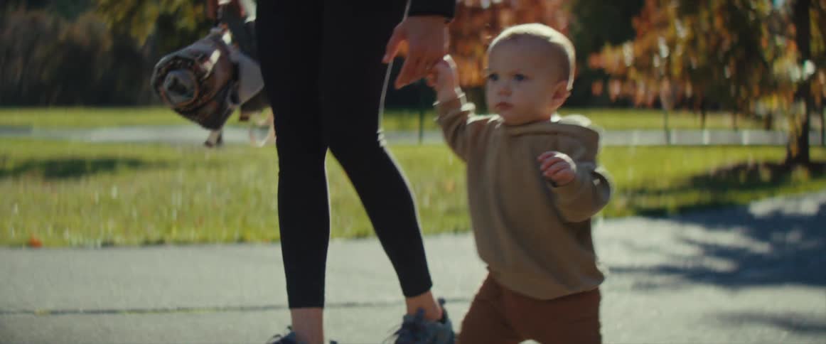 A little boy in a brown coat holding an adults hand walking along a path in a park with trees