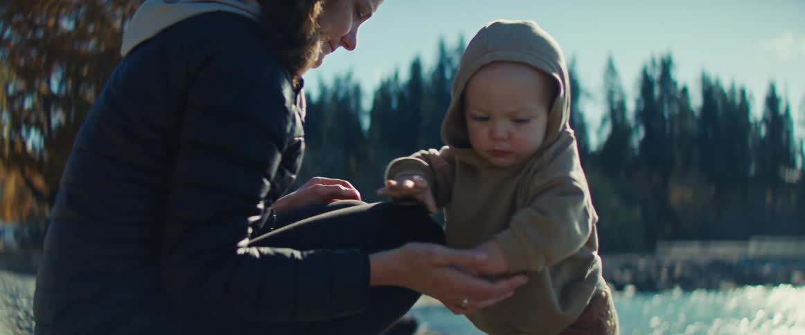A parent crouches down to help a bundledup toddler pull their hood up while standing outdoors near a body of water the scene is bright with a backdrop of tall pine trees suggesting a beautiful and brisk natural setting