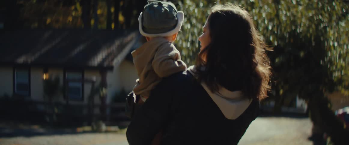 A parent carries a toddler in a cap through a sunlit area as the child drops their cap
