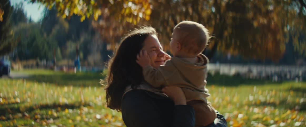A woman plays with her baby in a sunlit park surrounded by autumn leaves