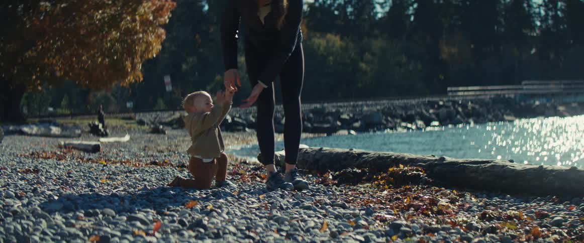 A mother and a young child are walking along a rocky beach or shoreline on a bright sunny daythe mother holds the childs hand as they walk with the shimmering water distant mountains and evergreen trees visible in the background